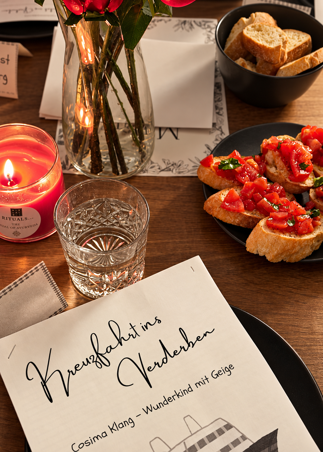 Gedeckter Tisch mit Kerze, Rosen und Bruschetta, davor ein Rollenheft von Kreuzfahrt ins Verderben